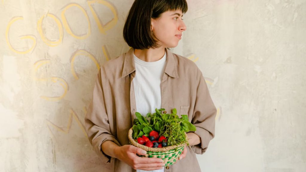 Woman standing with a basket of fresh greens and berries against a rustic wall background.