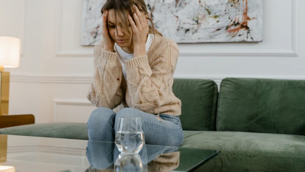 A woman sits on a green sofa looking stressed, holding her head with her hands.
