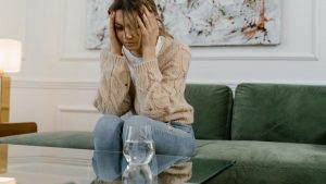 A woman sits on a green sofa looking stressed, holding her head with her hands.